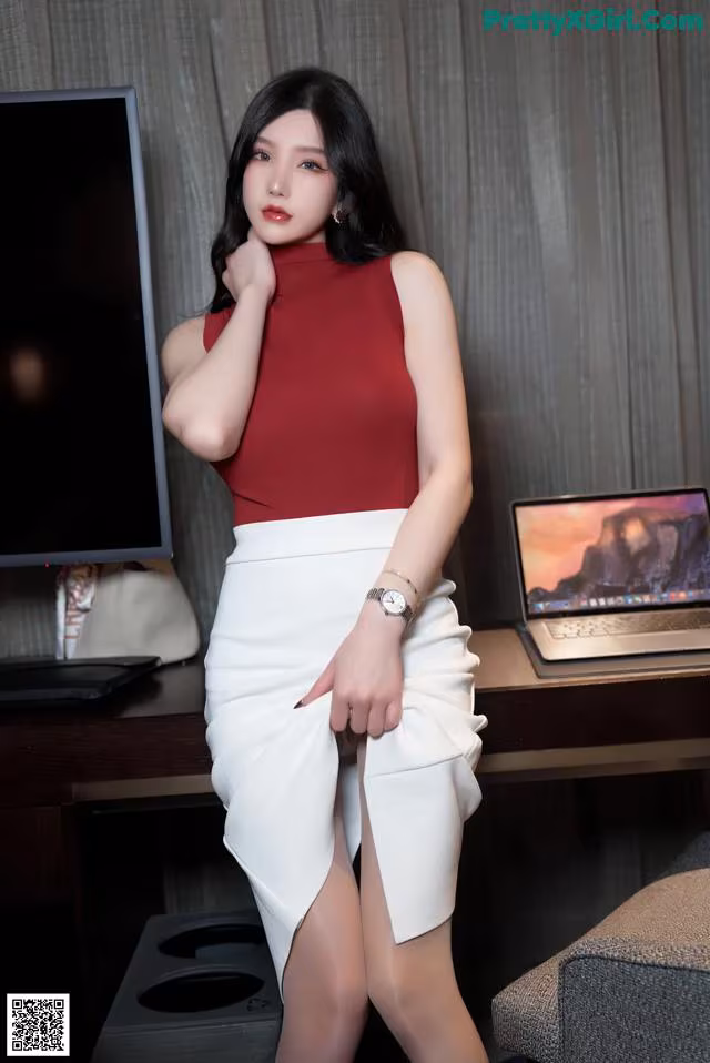A woman in a red top and white skirt posing in front of a computer.