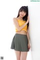 A woman in a yellow top and green skirt leaning against a wall.